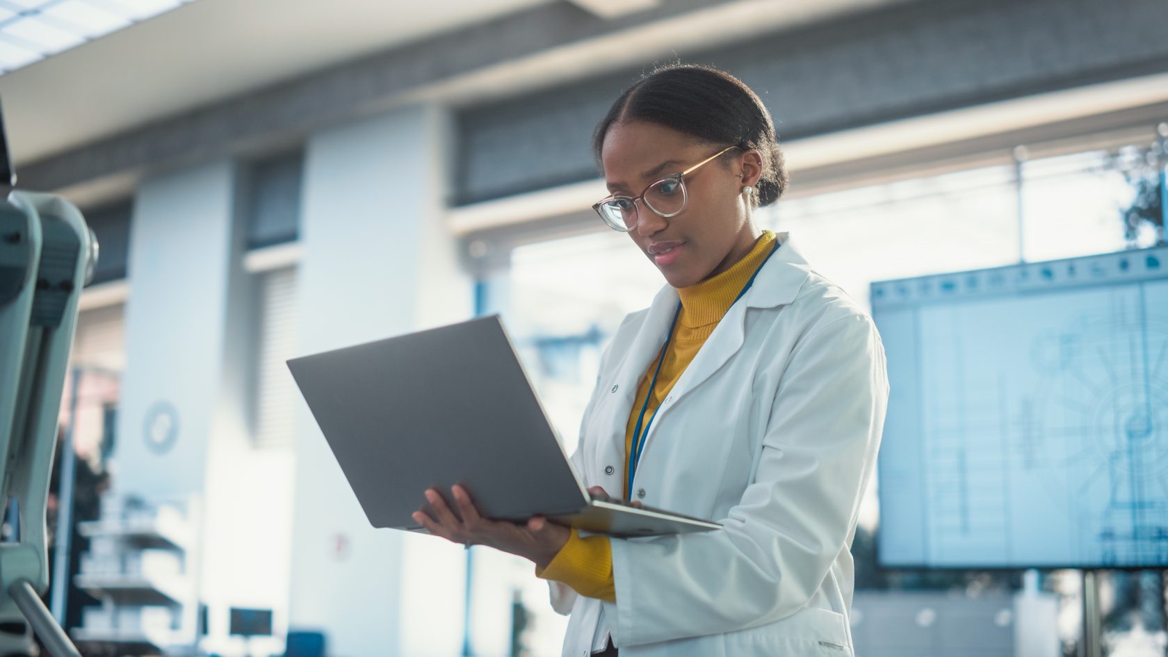 Female engineer using a laptop in a technical lab environment with robotic equipment and engineering diagrams visible in the background