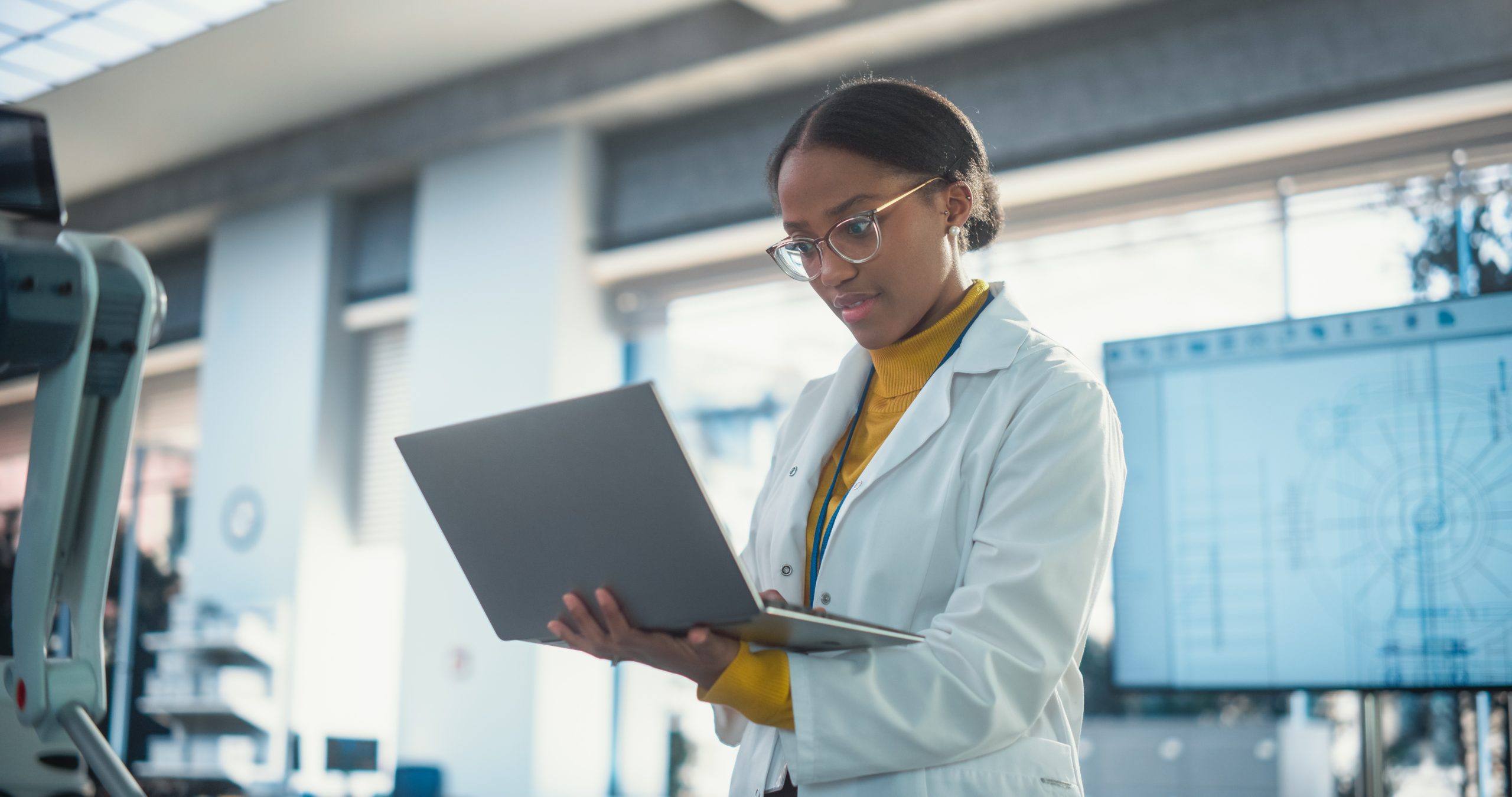 Female engineer using a laptop in a technical lab environment with robotic equipment and engineering diagrams visible in the background